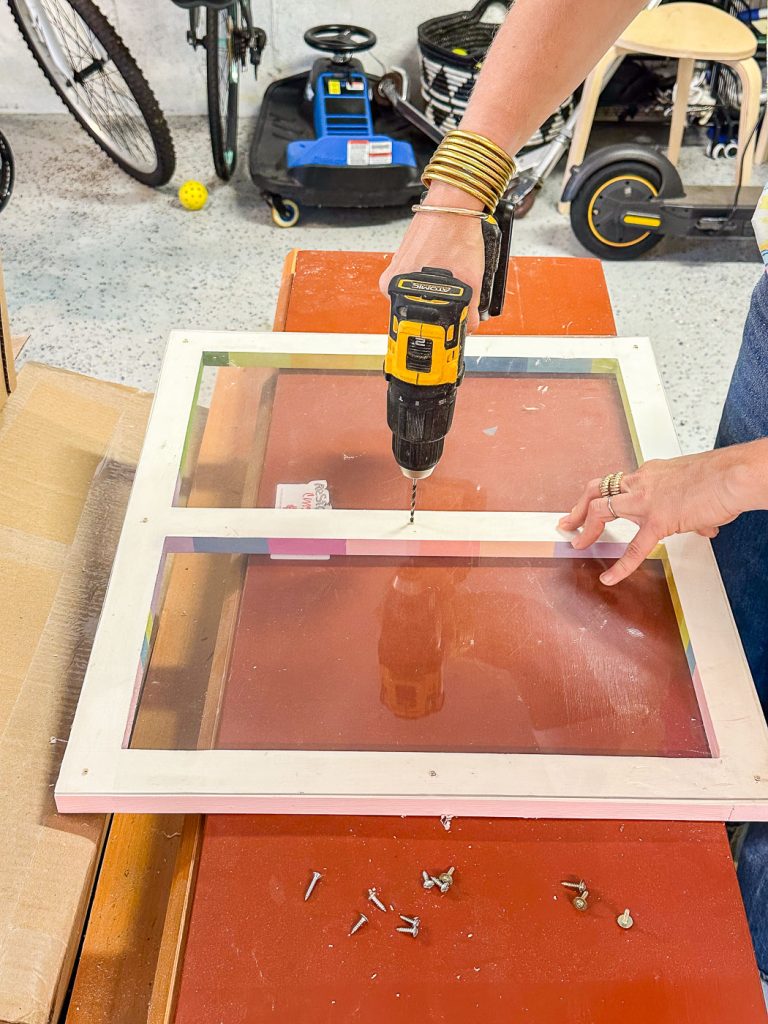 A person wearing bangles uses an electric drill to screw a sheet of acrylic onto a door frame on a table. Various tools, screws, and bicycles are visible in the background. The table is cluttered with different materials.