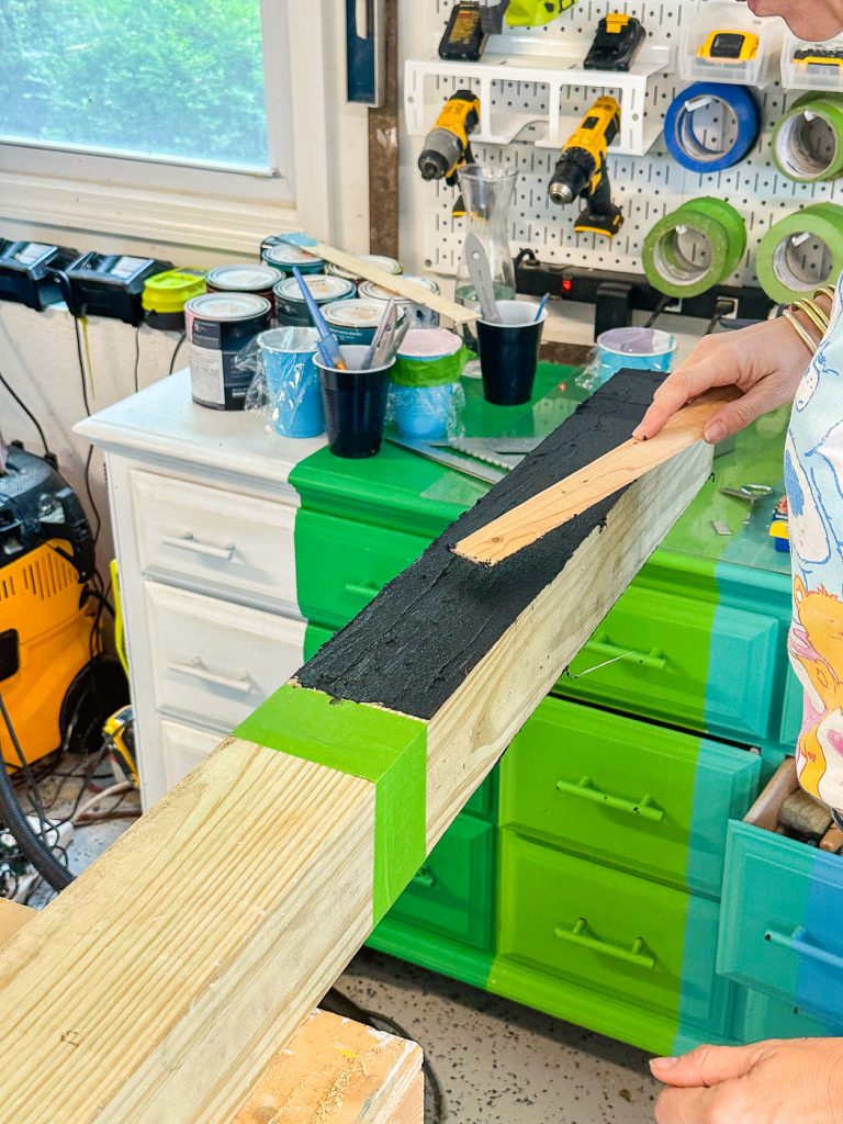 Person applying black tar to a wooden plank in a workshop, surrounded by various tools, paint cans, and equipment. The workspace includes a brightly painted green and blue cabinet and a window. Green painter's tape marks off sections on the plank.