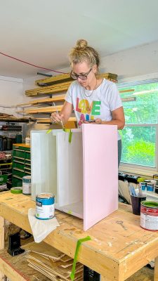 A woman with blonde hair in a bun, wearing glasses and a white t-shirt with "LOVE" written in colorful letters, is standing on a wooden table painting the exterior of a box pink. The setting appears to be a workshop with various tools and materials around.