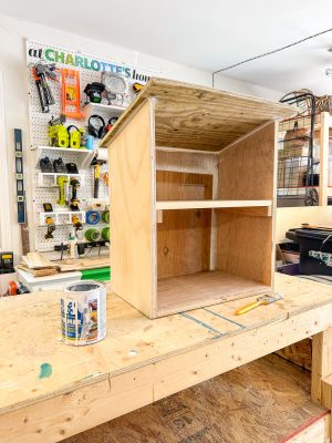 A wooden cabinet is being constructed in a workshop, featuring one interior shelf and an unfinished surface. Tools and materials, including a can of wood filler, primer, and a putty knife, are placed on the workbench. The background shows a pegboard with various tools hanging on it.