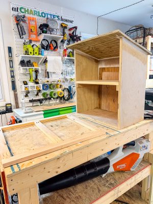 A workshop features a partially constructed wooden cabinet on a large workbench. Various tools, including drills, paint rollers, and levelers, hang on a pegboard against the wall. A Stihl power tool is visible beneath the workbench.