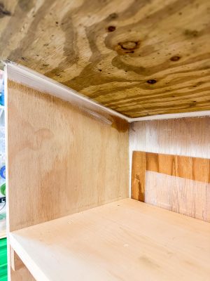 Close-up of a partially constructed wooden shelf unit. The raw plywood features visible wood grain and edges that appear to be partially coated with wood filler or sealant, along with some caulk in the joints. Ambient light illuminates the interior of the shelf.