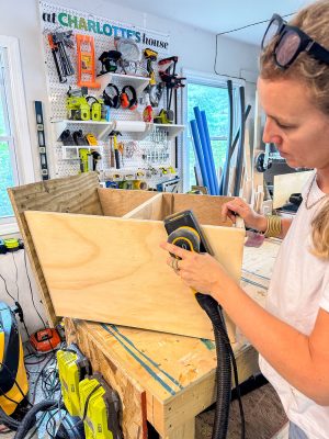 A person wearing sunglasses on their head is sanding a wooden structure with an electric sander in a well-organized workshop. The background displays various tools hanging on a pegboard and multiple windows, with natural light illuminating the workspace.