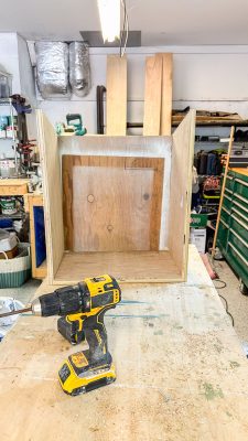 A wooden box is being assembled on a workbench in a cluttered workshop. A yellow and black cordless drill is placed in front of the box. Various tools, wood pieces, and equipment are scattered around the workspace.