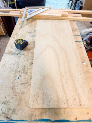 A woodworking workspace showing a rectangular plywood board on a workbench. A measuring tape and a metal square tool are also on the bench. Additional wooden strips are placed near the back of the bench. The area appears organized and ready for a project.