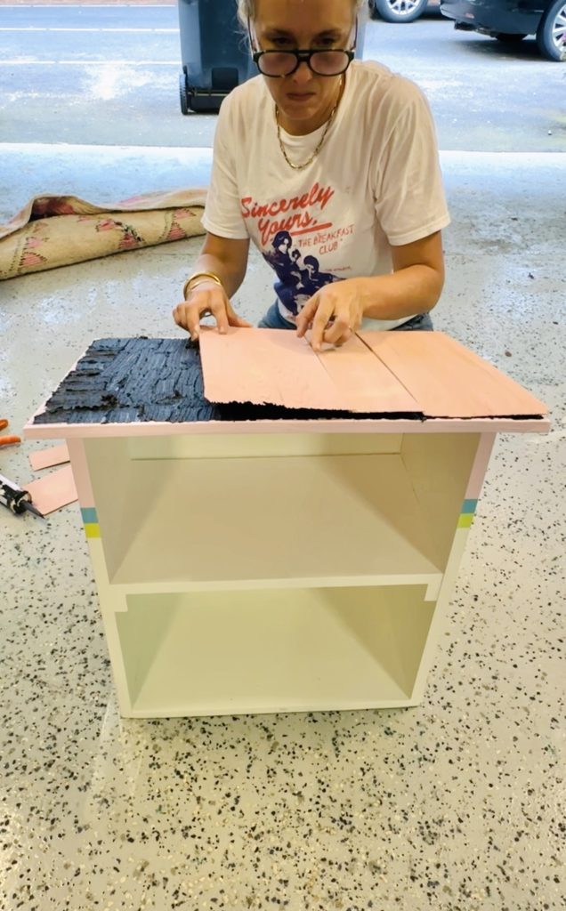 A person wearing glasses and a white printed t-shirt works on crafting, applying pink material to the top of a small white shelf unit. Tools and craft supplies are scattered around on the spotted tile floor in a room with a rolled-up rug in the background.