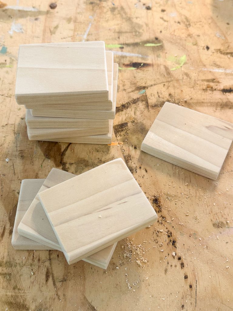 A stack of small, rectangular wooden pieces sits on a wooden workbench. Additional pieces are scattered nearby. The workbench shows signs of use with wood shavings, sawdust, and paint splatters.