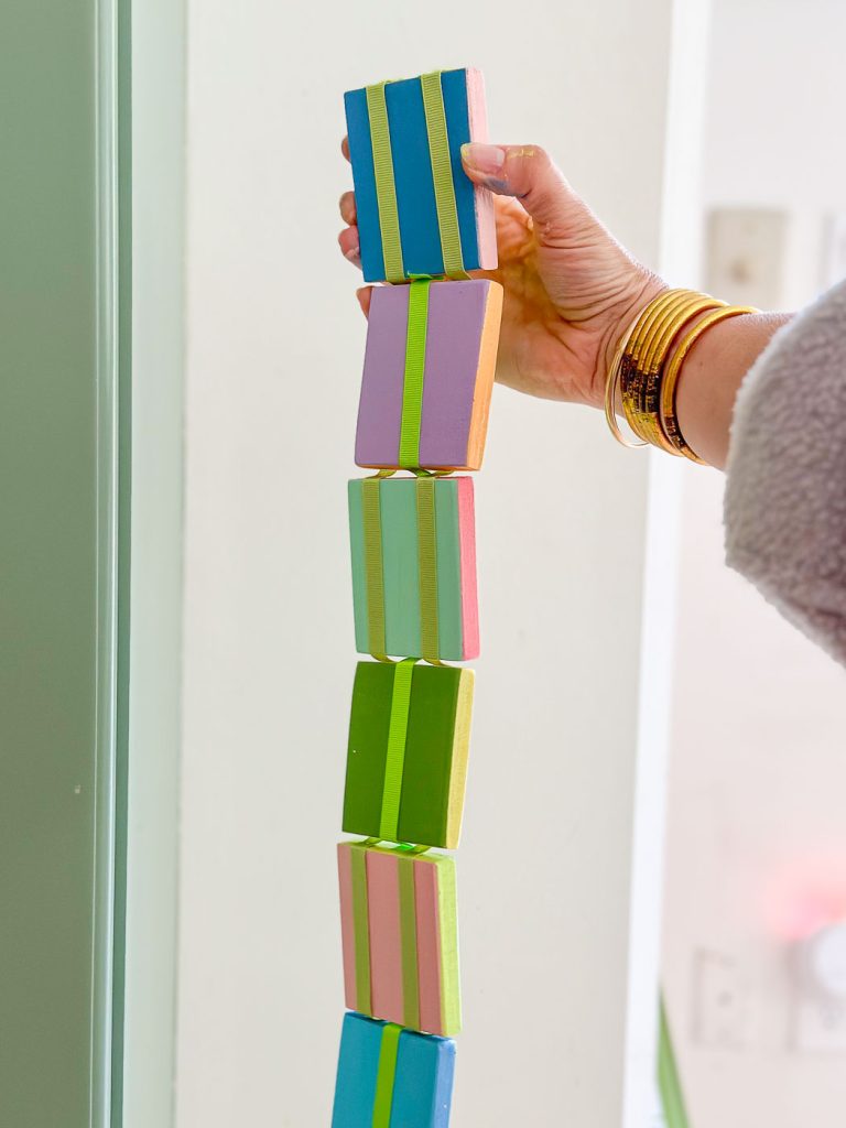 A hand wearing gold bangles is holding a vertical stack of colorful rectangular blocks. The blocks are in pairs of blue and pink, green and lime, pink and blue, and orange and green, and appear to be carefully balanced.