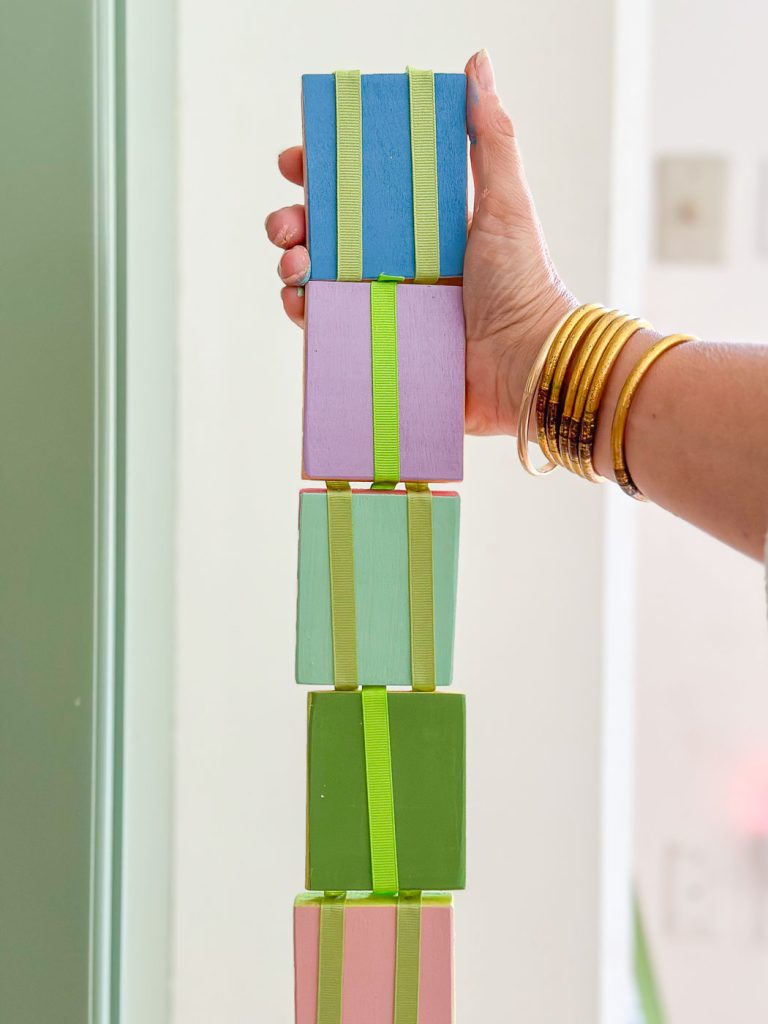 A person wearing multiple gold bangles holds up a vertical, rainbow-colored stack of small wooden blocks connected by green ribbons. The blocks are painted in blue, purple, green, and pink, and the background appears light and out of focus.