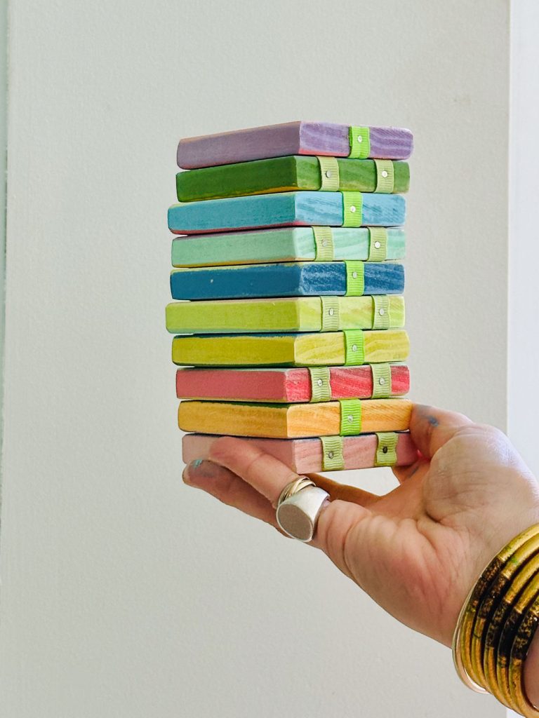 Close-up of a hand holding a stack of nine colorful, rectangular wooden blocks, each bound by two green bands. The blocks have various colors like purple, red, blue, and yellow. The hand is adorned with multiple gold bangles and a large silver ring.