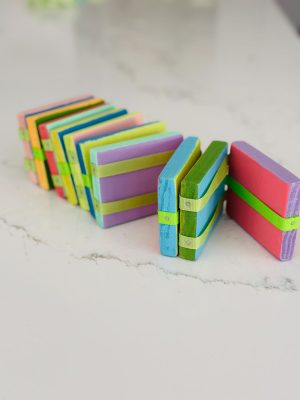 A colorful stack of rectangular wooden blocks, painted in various shades of blue, green, pink, and purple, are tied together with green ribbons. The blocks are positioned in a zigzag pattern on a white marble surface.