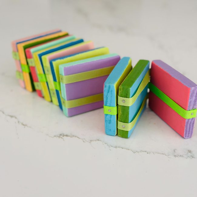 A colorful stack of rectangular wooden blocks, painted in various shades of blue, green, pink, and purple, are tied together with green ribbons. The blocks are positioned in a zigzag pattern on a white marble surface.