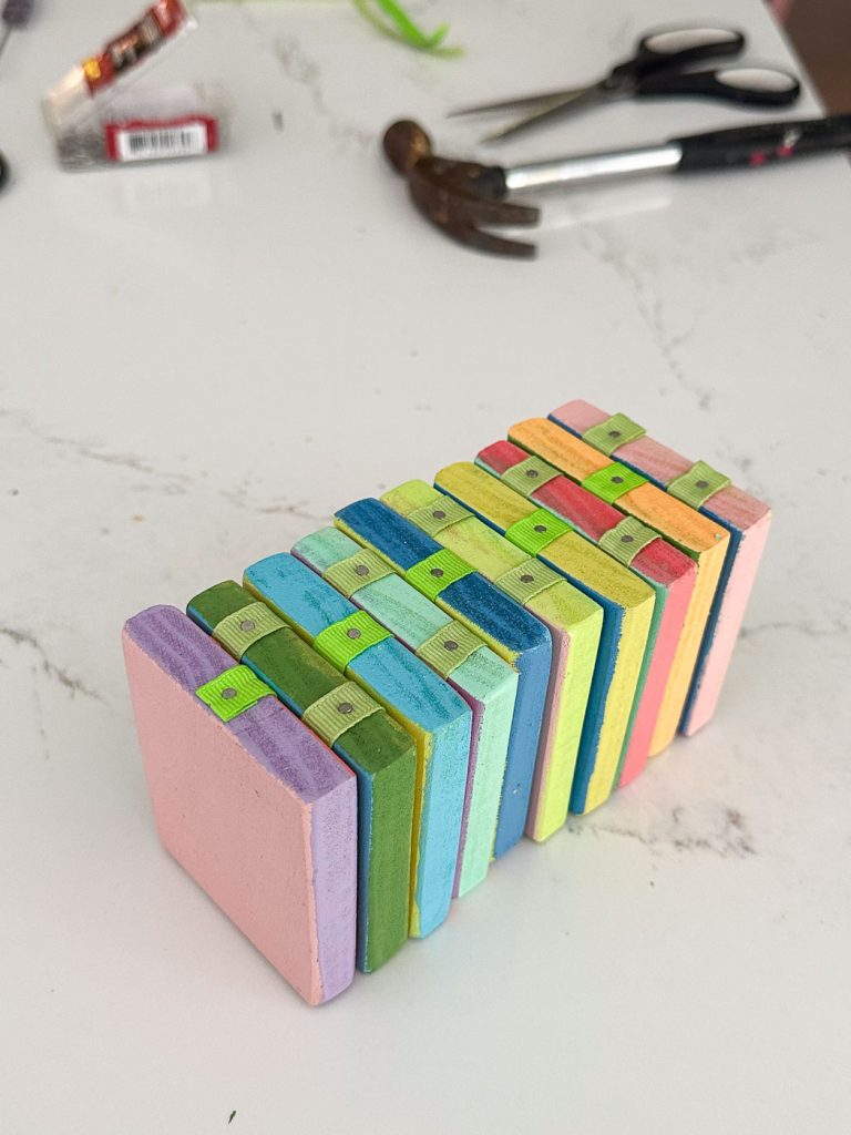 A set of colorful wooden blocks stacked in a zigzag pattern on a white marble surface. In the background, blurred items including a pair of scissors, a hammer, and a package of batteries can be seen.