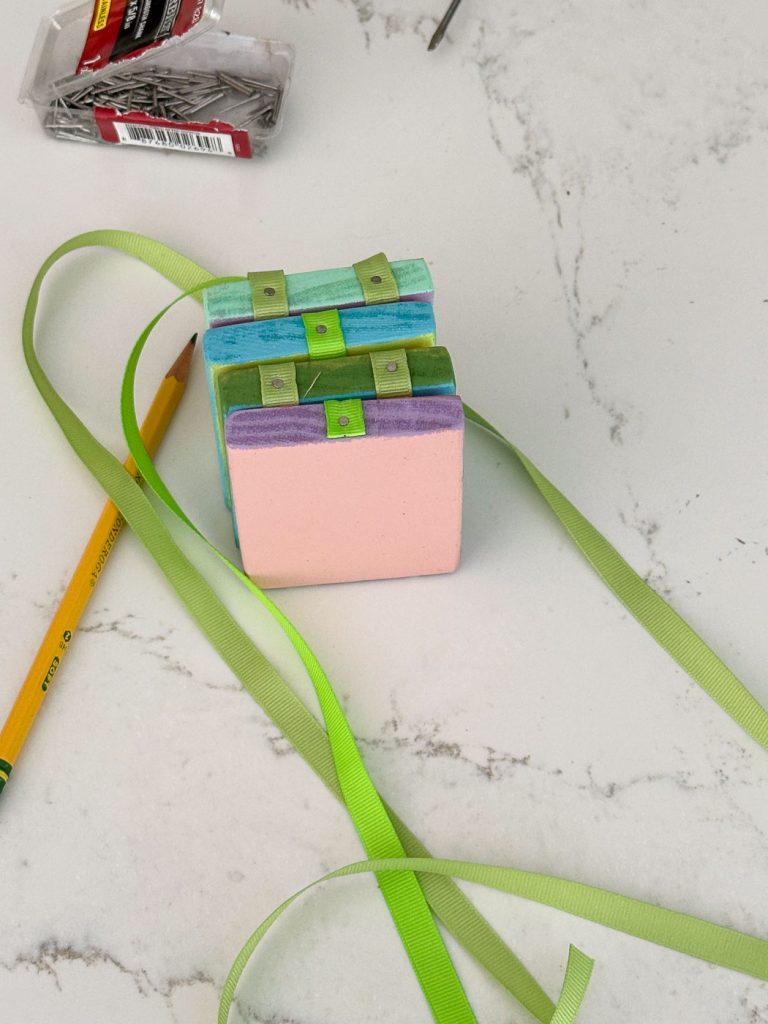 Two small, pastel-colored wooden blocks are stacked and tied together with a green ribbon. A yellow pencil lies nearby on a marble surface. In the background, there is an open box of nails.