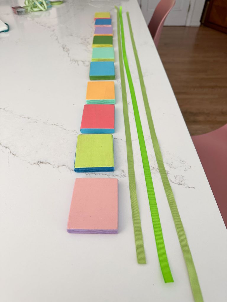 A row of colorful foam squares is aligned on a white marble surface, with each square being a different color (yellow, green, blue, pink, and orange). To the right of the squares, four green ribbons are laid out parallel to them.