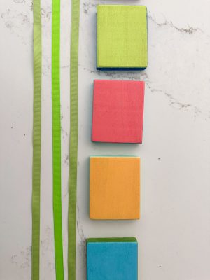 Four colored wooden blocks (green, red, yellow, and blue) are aligned vertically on a white marble surface with two parallel green ribbons to their left.