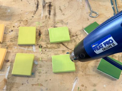 A workspace with six small, square pieces of wood, each painted in varying shades of yellow and green. A heat gun labeled "Heat Pro Deluxe II" is aimed at the pieces. The wooden surface has paint splatters and a pair of pliers in the top right corner.