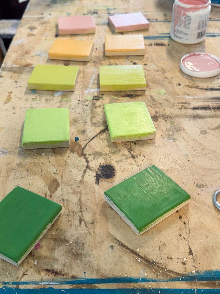 A wooden work surface displays small square wooden pieces in varying stages of being painted in shades of green, yellow, and peach. An open can of pink paint sits nearby. The table has visible paint splatters and marks from previous projects.