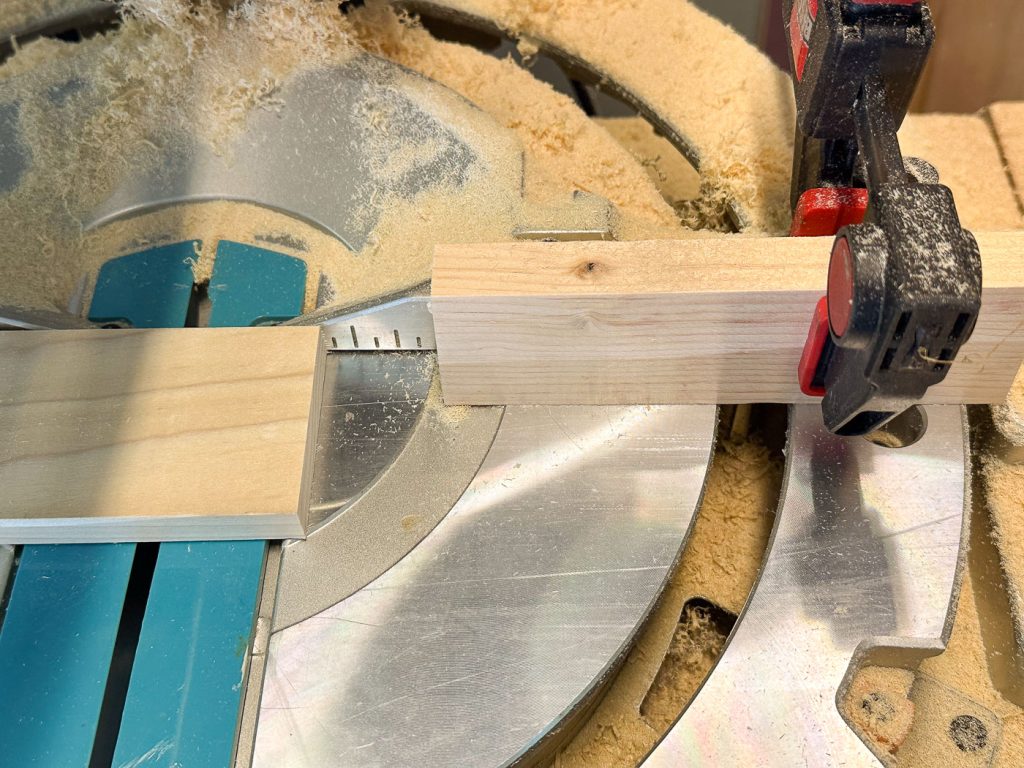 A circular saw with a wooden plank clamped in place, ready to be cut. Sawdust is scattered around the cutting area, and the saw's blade is partially visible. The plank is aligned with the miter gauge for precise cutting.