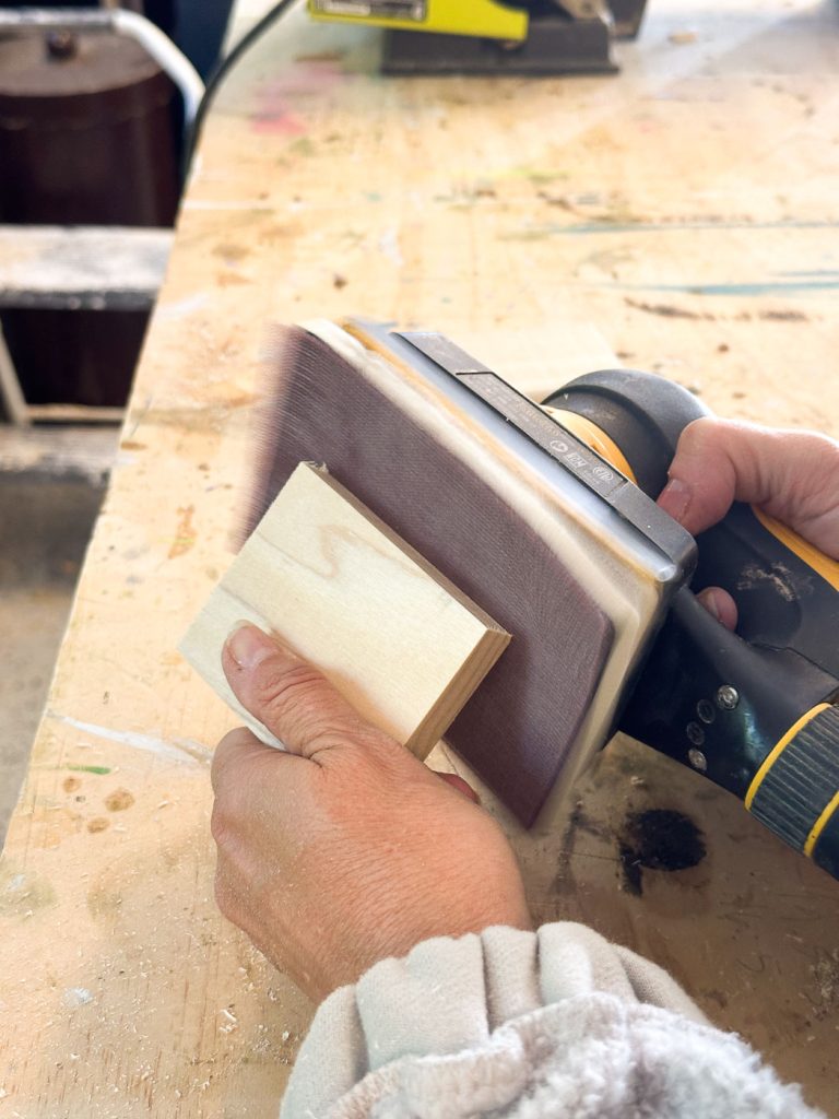 A person using a power sander to smooth the surface of a small wooden block on a workbench. The workspace is cluttered, and the person's hand is gripping the sander firmly while holding the block in place with the other hand.