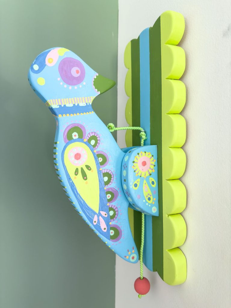 A wall-mounted blue wooden bird with colorful decorative patterns, including green, yellow, purple, and pink accents. The bird is attached to a vibrant yellow and green striped base, with a red bead hanging from a green string beneath it.