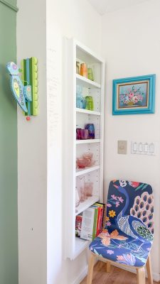 A corner setup featuring a quirky, colorful interior design with a chair adorned with vibrant, playful patterns. A narrow white shelf holds an array of colorful glassware. A decorative wall hook shaped like birds is on the left wall, and a framed floral painting is on the right.