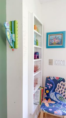 A corner of a room with a floral-themed chair, a small white bookshelf filled with colorful glassware, and a wall-mounted blue and green wooden bird decoration. A framed painting of flowers hangs on the wall above the chair.