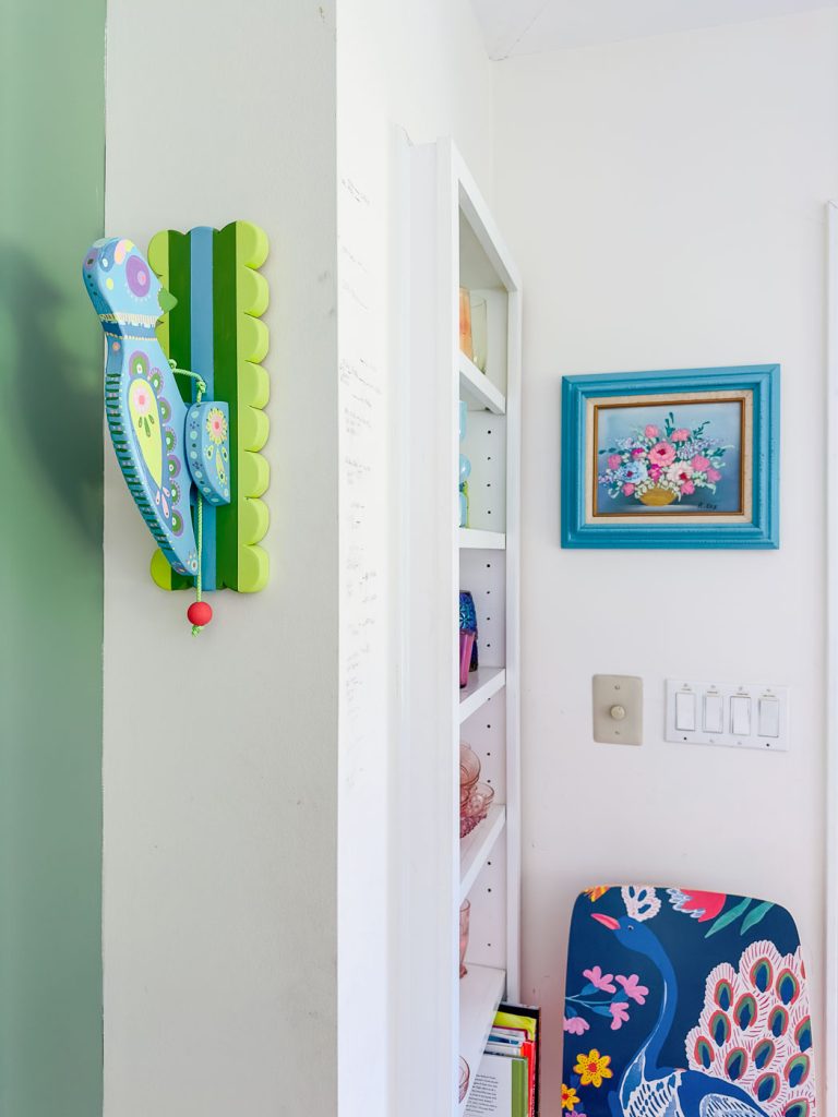 A colorful room features a vibrant bird-shaped wall decoration on a green mounted bracket, a turquoise framed painting of flowers, white shelves filled with various items, and a chair adorned with a peacock design. The wall also includes a light switch panel.