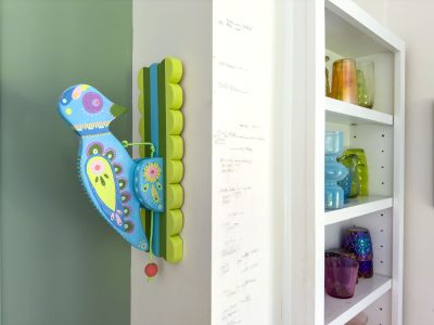 A colorful, decorative wooden bird is mounted on the corner of a wall with vibrant patterns. Nearby, a white shelf holds various glass vases and jars in different colors and sizes. The wall next to the bird shows markings, likely for measuring height.