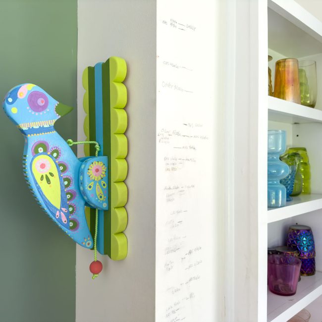 A colorful, decorative wooden bird is mounted on the corner of a wall with vibrant patterns. Nearby, a white shelf holds various glass vases and jars in different colors and sizes. The wall next to the bird shows markings, likely for measuring height.