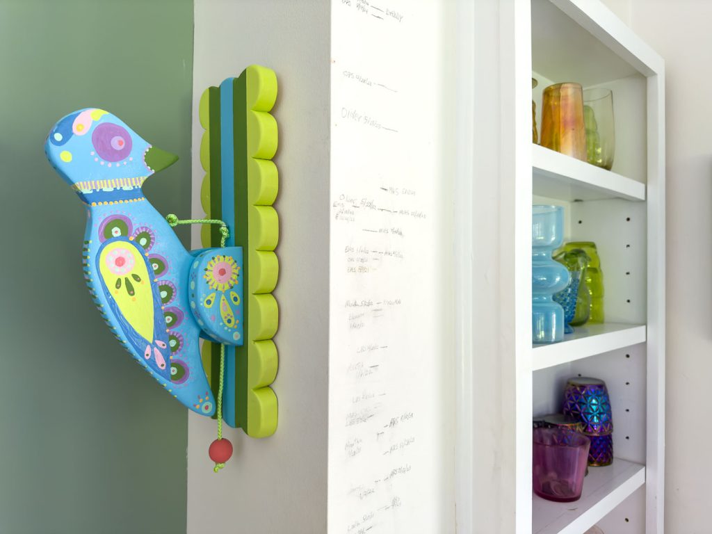 A whimsical, colorful bird decoration with intricate designs is mounted on a wall. The bird features shades of blue, green, and pink. To the right, there is a white shelf filled with various colorful glass jars and vases. The wall appears to have handwritten notes.