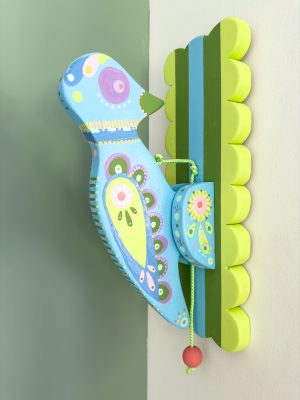 A colorful wooden bird decoration is mounted on a wall. The bird is painted in shades of blue with green, yellow, and pink patterns. It is connected to a vibrant green and blue striped wooden base with wavy edges, holding a small orange bead on a string.