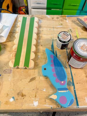 A cluttered workbench with a wooden item partially painted green on the left and a blue, bird-shaped wooden piece with pink accents on the right. Several open paint cans and brushes are scattered around, with paint splatters visible on the table.