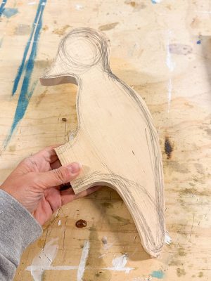 A hand holding a partially cut wooden piece with an outline sketch of a bird on it, resting on a wooden workspace scattered with pencil marks and light stains. The bird-shaped outline features a distinct body and head with circular eye detail.