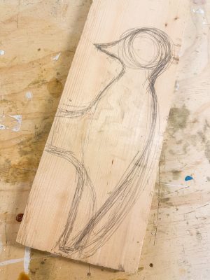 A wooden plank with a pencil sketch of a bird on it, depicting the outline of the bird's head and body. The plank is placed on a slightly stained workbench.