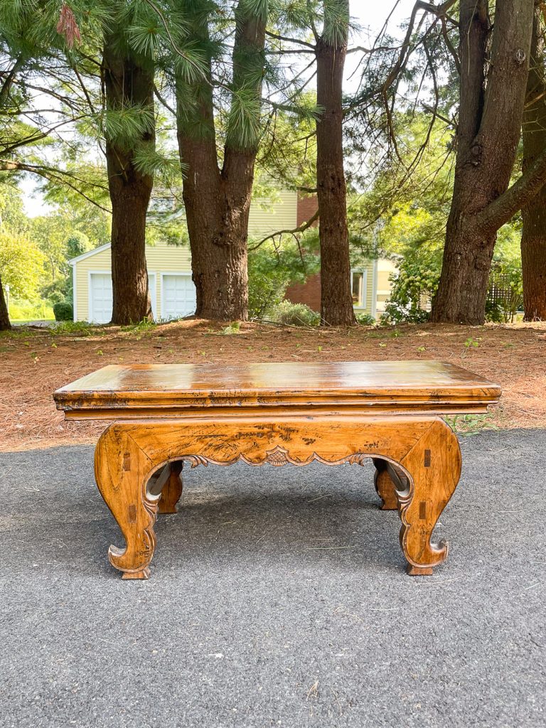 A rustic wooden table with intricate carvings sits on a paved path. It's surrounded by tall trees and greenery. In the background, a yellow house with white trim is partially visible.