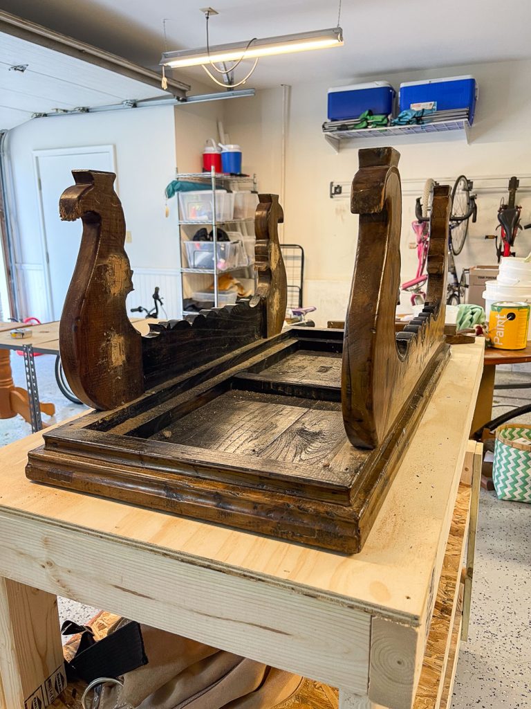 An upside-down wooden table with ornate legs sits on a workbench in a garage. Shelving, storage bins, and bicycles are visible in the background.