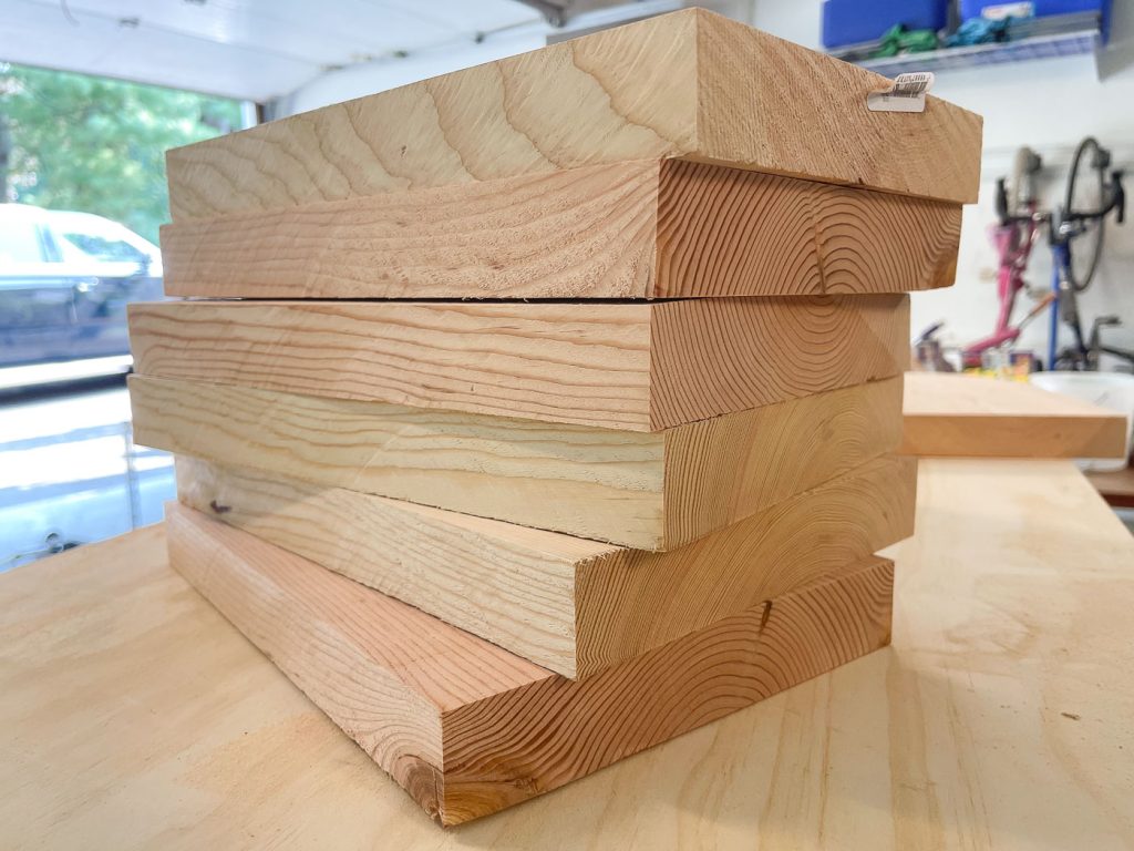 A stack of six rectangular lumber pieces on a wooden table in a garage. Natural wood grain is visible. The background includes a parked car, a window with greenery outside, and some hanging tools and shelves.