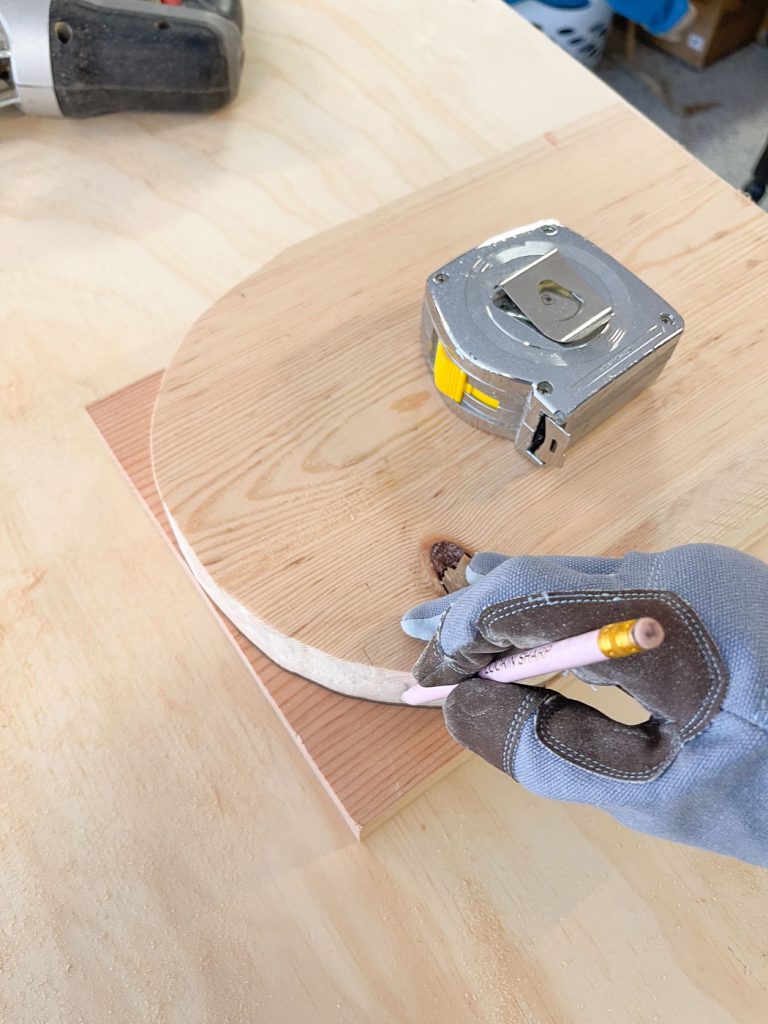 A gloved hand holds a pencil, tracing a curved line on a piece of wood placed on a table. A tape measure rests on top of the wood. The scene suggests woodworking or a DIY project in progress.