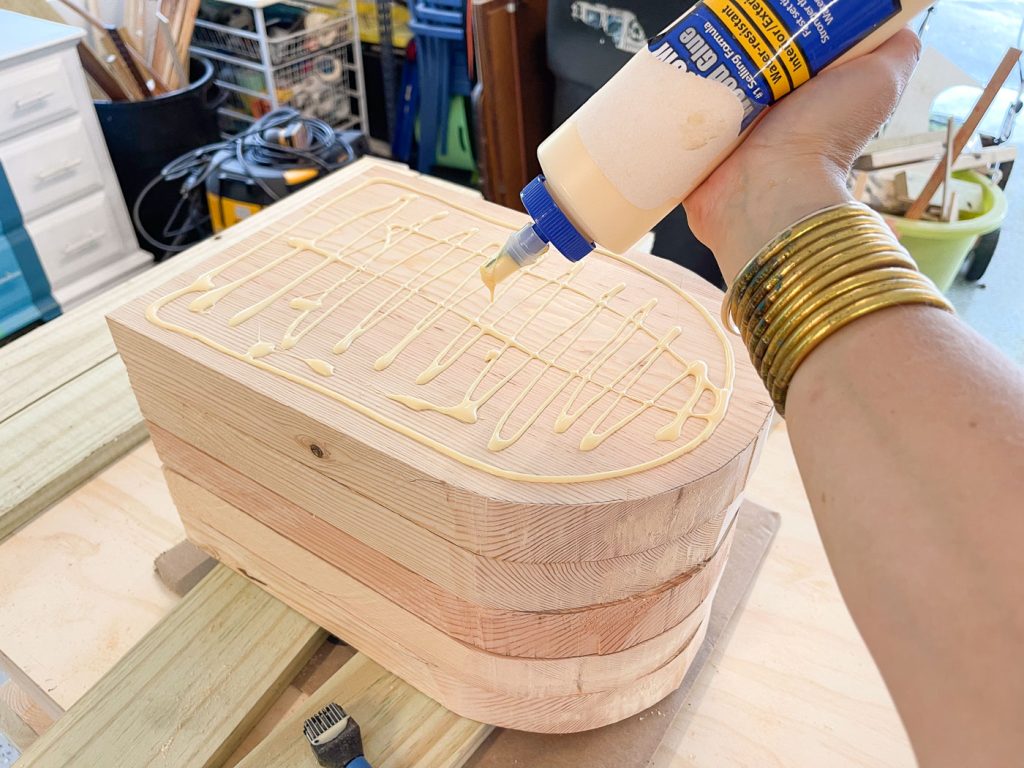 A person applies wood glue to a stack of curved wooden pieces on a workbench. The wood is neatly arranged, and there's a brush nearby. The setting appears to be a workshop with various tools and materials visible in the background.