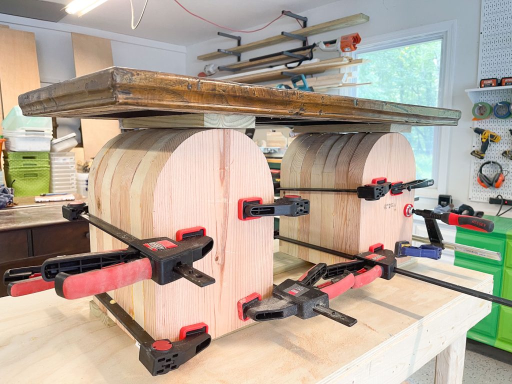 A woodworking project in progress, featuring two large wooden forms held together with multiple black and red clamps. A wooden plank rests on top. Various tools and supplies are visible in the background of a workshop.