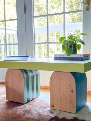 A modern, colorful table with wooden and green painted arches supports a white rectangular tabletop. It is adorned with a small plant and a stack of books. The table is positioned near a bright window with a view of greenery outside.
