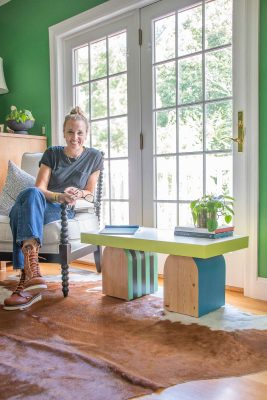 A person with hair in a bun sits on a black chair, smiling. They're next to a colorful coffee table with a potted plant and books. The room has green walls and a cowhide rug, with large windows in the background letting in natural light.