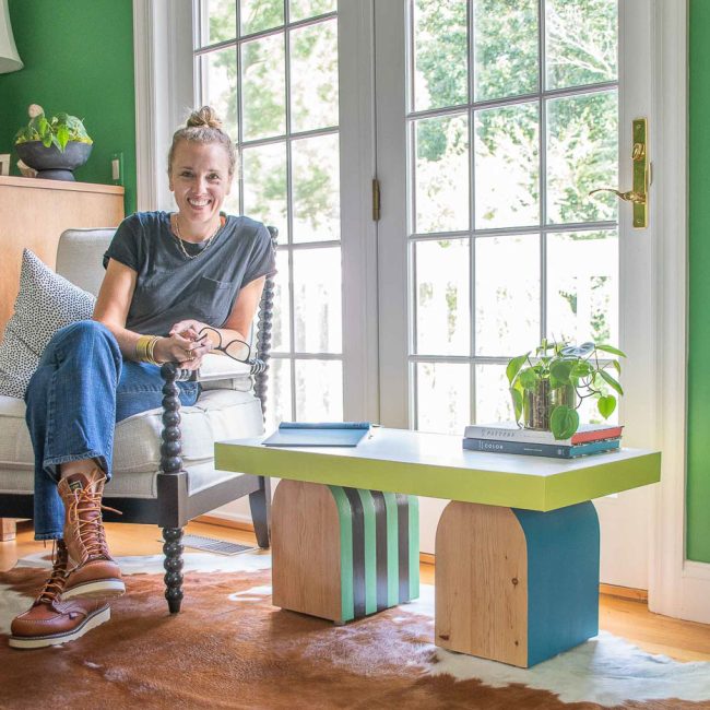 A person with hair in a bun sits on a black chair, smiling. They're next to a colorful coffee table with a potted plant and books. The room has green walls and a cowhide rug, with large windows in the background letting in natural light.