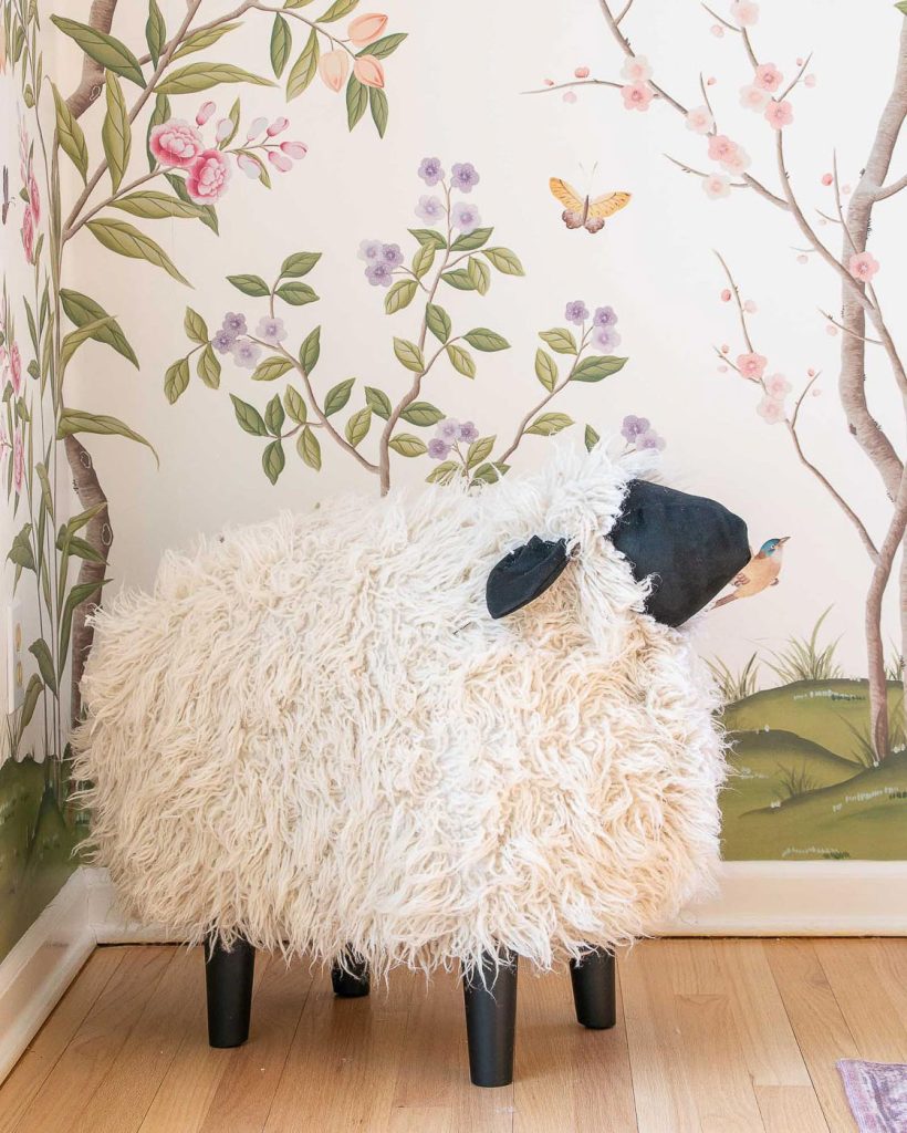 A fluffy sheep-shaped stool with black legs is placed in a corner, set against a backdrop of a floral-patterned wall with illustrated trees, flowers, and a butterfly. The room has light wooden flooring.