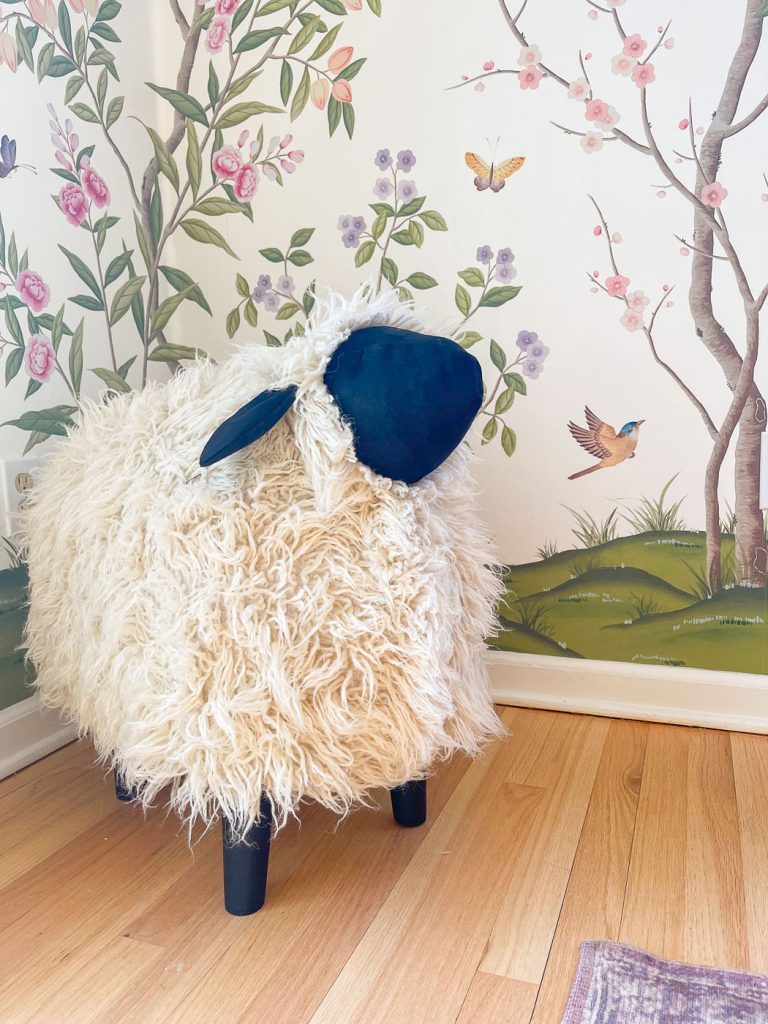 A fluffy sheep-shaped ottoman with black legs and ears stands on a wooden floor. The wall behind features a colorful mural with birds, butterflies, and flowers.