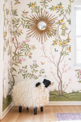 A fluffy sheep-shaped stool stands in a corner with floral wallpaper featuring birds and butterflies. A sunburst mirror hangs above, reflecting the room. The wooden floor complements the cozy, whimsical decor.