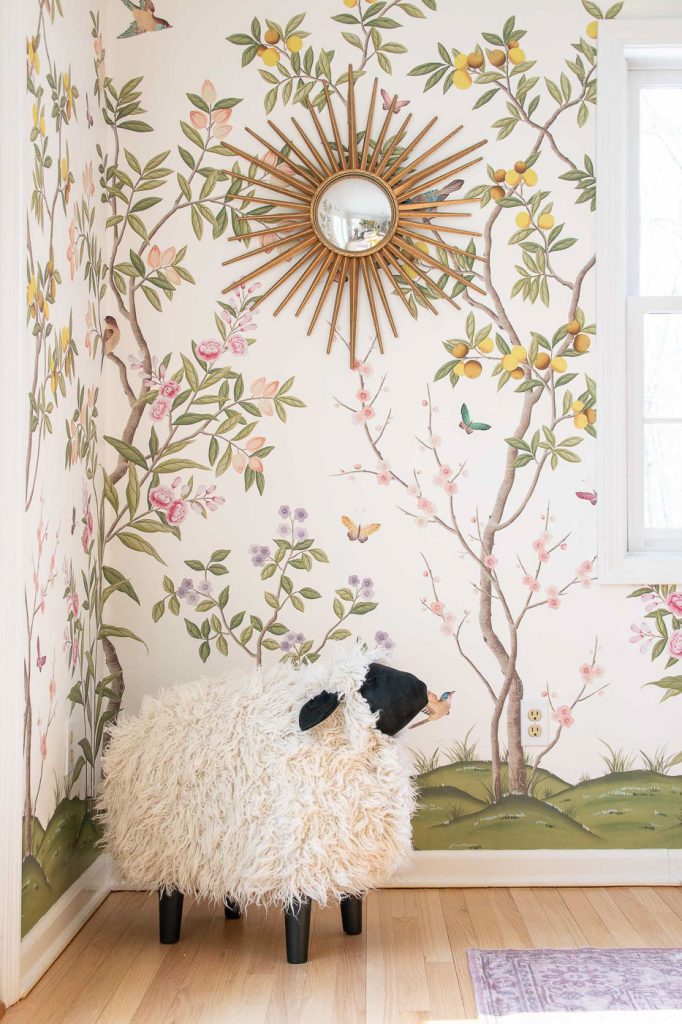 A fluffy sheep-shaped stool stands in a corner with floral wallpaper featuring birds and butterflies. A sunburst mirror hangs above, reflecting the room. The wooden floor complements the cozy, whimsical decor.