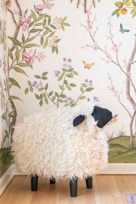 A fluffy sheep-shaped stool with black legs and face stands in a corner. The walls are decorated with a colorful floral and butterfly mural on a light background. The floor is wooden.