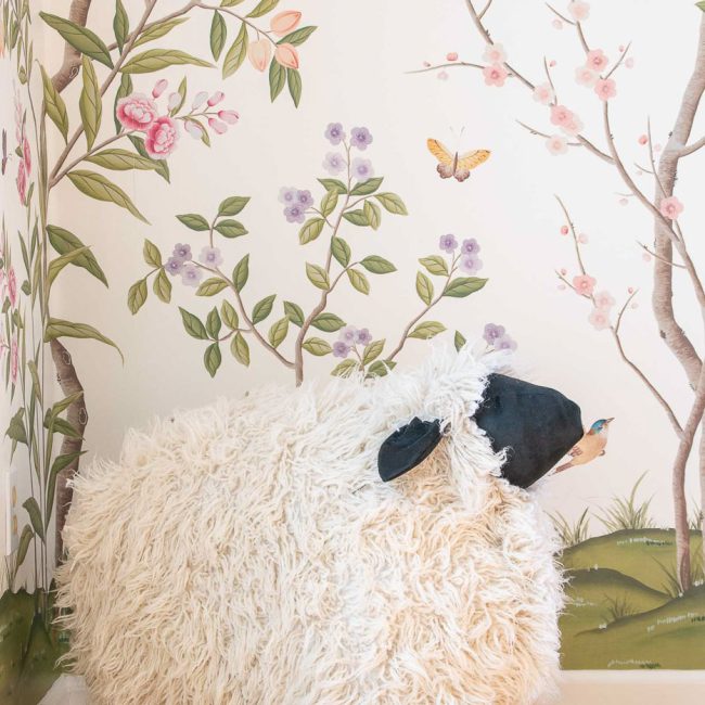 A fluffy sheep-shaped stool with black legs and face stands in a corner. The walls are decorated with a colorful floral and butterfly mural on a light background. The floor is wooden.